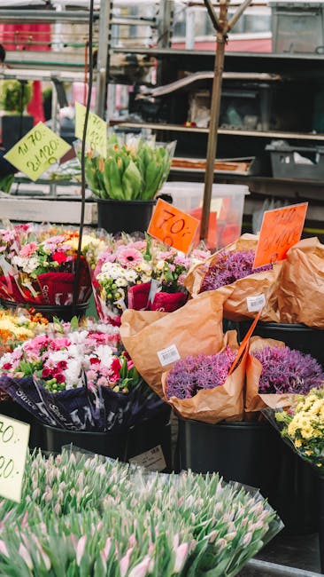 A vibrant outdoor market display featuring a variety of fresh flowers arranged in black plastic buckets and wrapped bouquets. In the foreground, there are pink tulips with closed petals, and behind th