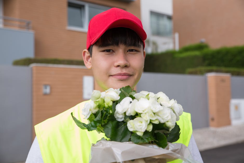 A young male flower delivery courier wearing a red cap and a high-visibility yellow vest is standing outdoors in front of modern residential buildings. He is smiling slightly and holding a bouquet of 