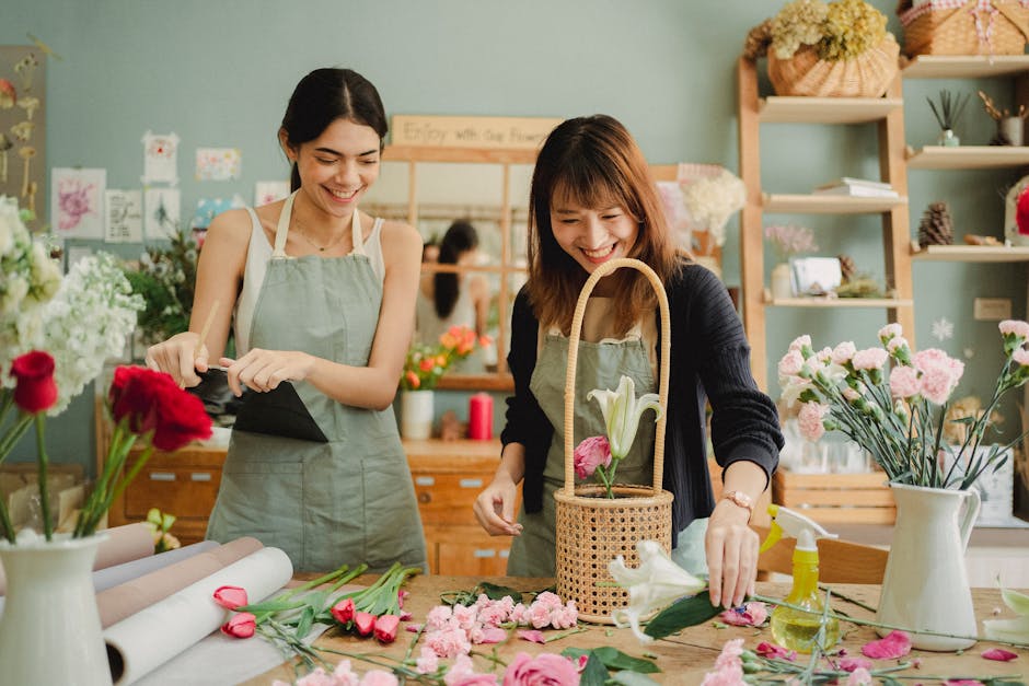 Two women are working together in a bright floral shop, arranging a bouquet of fresh flowers. The woman on the left has dark hair tied back and is wearing a light green apron, holding a black wrapper