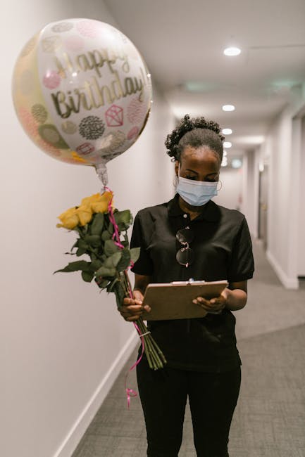 A florist in black attire and a face mask stands in a well-lit corridor, holding a bouquet of yellow roses wrapped in green foliage and pink ribbon. She also carries a round foil balloon with colorful
