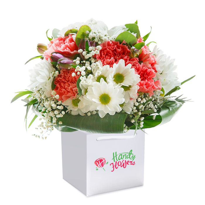 Elegance and vibrancy combine in this striking arrangement, set upright in a clean white gift bag and ready for same-day delivery across Harold Hill. The bouquet unfolds with an abundance of coral, peach, and rich scarlet-red carnations, each bloom distinguished by plush, textured petals that impart an inviting depth and celebration. Tucked among these are clusters of snow-white daisies with vivid yellow-green centers, bringing lively contrast and a glimpse of early spring on Harold Court Road. Sprigs of delicate white baby's breath add a touch of ethereal softness, drifting gently above the more robust blooms. A few closed buds, tinged with purple and hidden in the foliage, promise unfolding beauty in the days ahead. Broad, glossy green leaves form a lush collar at the base, cradling the arrangement with polished structure and zest-a testament to the thoughtful care of Harold Hill flower experts. The bouquet is meticulously arranged into a well-rounded, abundant shape, and its crisp presentation in a white gift bag with sturdy handles sets the tone for memorable gifting-a perfect gesture for any anniversary, milestone, or heartfelt surprise within the neighbourhood. The product image, set against a flawless white backdrop, highlights the freshness, quality, and artistic composition that distinguish the local florist's craft.