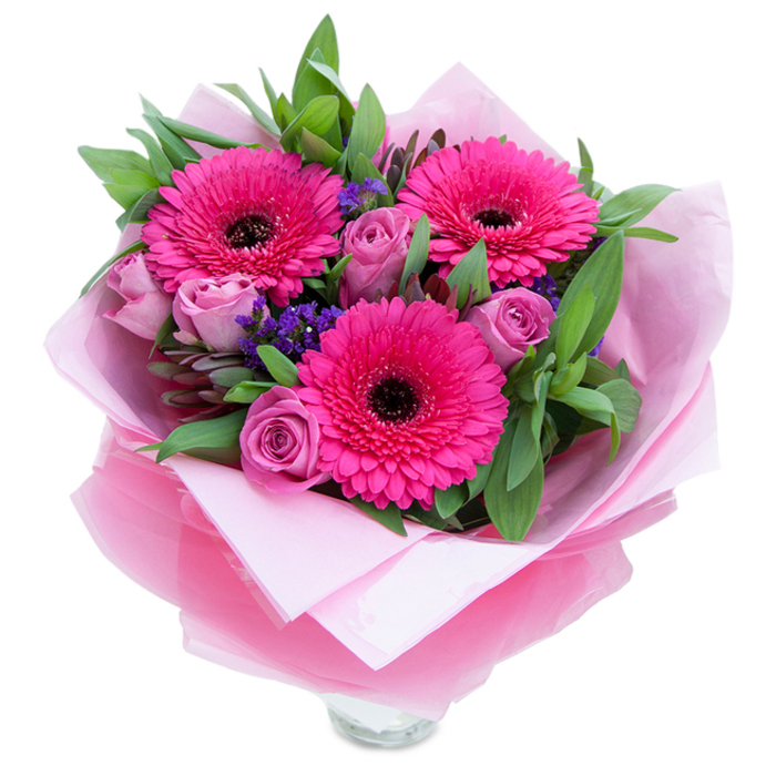 I see this bouquet as a blush-hued embrace captured against a plain white surface: three vibrant fuchsia Gerbera daisies take centre stage, their concentric petals unfurling around dark, espresso-coloured centres that add dramatic contrast and visual depth. Nestled close by are roses in a delicate lavender-pink or dusty rose shade, each bloom still showing the tight, layered curls of a bud that promises to open further and release a soft floral perfume. Interspersed between these are sprays of tiny purple statice or limonium, lending an airy, meadow-like texture and a cool tonal counterpoint to the warmth of the pinks. The supporting greenery-broad leaves that read as salal or eucalyptus-envelops the composition, giving it fullness and a subtly crisp, botanical scent. The bouquet is lovingly wrapped: an inner sheet of soft, matte pale pink tissue creates a gentle cushion, while an outer veil of slightly crinkled, translucent fuchsia-pink cellophane imparts a delicate shimmer and reinforces the colour story; the folds are artfully arranged to present the stems and blooms with care. At the base, the rim of a clear glass vase glints through, suggesting this arrangement is ready to brighten a living room table or hallway console in Harold Hill or a neighbouring street. The overall mood is celebratory yet tender-suitable for Mother's Day, a birthday, an anniversary, or a simple 'thinking of you'-crafted to uplift with both colour and thoughtful florist technique.