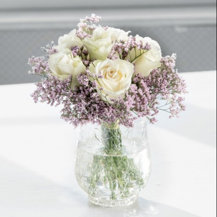 A carefully composed bouquet of creamy white roses and pale lavender statice stands proudly in a clear glass vase, bathed in gentle daylight that feels typical of a calm morning in Harold Hill. The arrangement is full and rounded, almost perfectly dome-shaped, with approximately a dozen white roses forming the main structure. Each rose is at that ideal stage of opening, where the outer petals are just beginning to curl back, revealing a softly layered centre. The petals themselves are rich and velvety, their tone more ivory than bright white, which lends the bouquet a warm, romantic glow. Threaded through and encircling these central blooms are plentiful sprays of light lilac statice. The statice is composed of tiny, clustered flowers on fine, branching stems, adding a feathery, cloud-like frame around the substantial rose heads. Their subtle purple hue introduces a gentle contrast that remains soothing and refined, rather than bold. All the stems are gathered into a tall, cylindrical glass vase, the kind often seen on clean, minimalist desks or console tables around Harold Court Road. Through the crystal-clear glass you can see the straight, green rose stems and the more delicate lines of the statice, all immersed in clear water that catches glints of light. The vase rests on a smooth, bright white surface, which bounces soft reflections back onto the lower blooms and gives a sense of space and cleanliness. The background is blurred, implying an indoor setting with a large window letting in natural light, perhaps in a modern Harold Hill flat or office. The overall impression is one of modern simplicity paired with classic charm, suitable for anniversary deliveries, peaceful sympathy tributes, or quiet moments of celebration at home.
