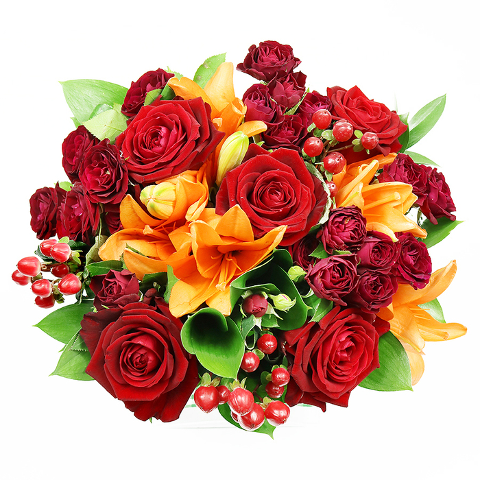 Captured from above on a pristine white field, this sumptuous, densely packed bouquet reads like an heirloom study in warm tones and tactile contrasts. Several sumptuous deep red roses dominate the composition, their velvety petal spirals opened wide to expose layered centres that convey timeless romance and a substantial, fragrant presence. Counterpointing the roses are bright orange Asiatic lilies, their trumpet forms and visible stamens introducing dynamic, architectural lines; some lilies are mothlike and fully spread, others are elegantly furled, suggesting movement and the promise of continued bloom. Interspersed throughout are clusters of deep burgundy spray roses whose miniature blooms provide rich shadow and a luxurious, velveteen texture. Scattered among blossoms, glossy red hypericum berries punctuate the palette with concentrated points of light and a cheerful, tactile contrast to the soft petals. Broad, glossy foliage-likely ruscus or salal-frames the bouquet and gives it a fresh, green backbone that lifts the warm hues. The top-down perspective emphasises the perfectly rounded silhouette and the careful, hand-arranged rhythm of blooms and negative space, while soft, diffuse lighting brings out both colour saturation and delicate petal detail without dramatic shadow. This arrangement reads as celebratory and heartfelt, ideal for anniversaries or seasonal gatherings, and would sit beautifully on a dining table in a Harold Hill home or be chosen to mark a meaningful moment in nearby Romford. A seasoned florist would note the balance of scale, scent, and texture-an opulent yet approachable composition.